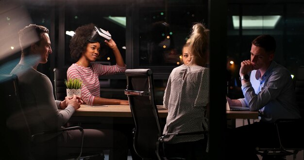 Team aziendale multietnico che utilizza cuffie per realtà virtuale nella riunione notturna dell'ufficio Gli sviluppatori si incontrano con il simulatore di realtà virtuale attorno al tavolo nell'ufficio creativo.