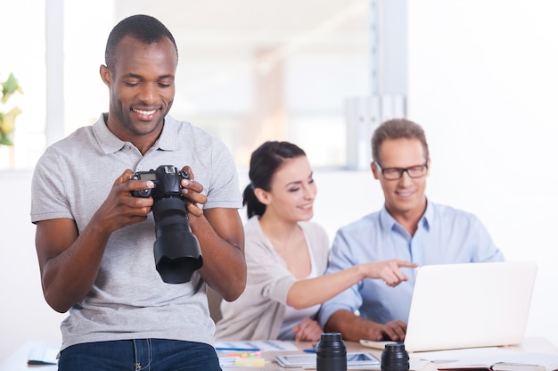 Team creativo al lavoro. Bel giovane africano che tiene la macchina fotografica e sorride mentre due persone lavorano sullo sfondo