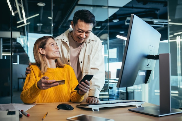 Team creativo di uomini e donne asiatici che acquistano online seduti a un tavolo scegliendo un regalo insieme gioendo e sorridendo utilizzando uno smartphone e una carta di credito