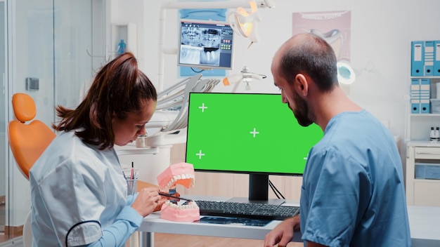 Team di igiene orale che analizza la disposizione dei denti durante l'utilizzo di un computer con schermo verde orizzontale sulla scrivania. Dentista e assistente dentale che lavorano con strumenti e attrezzature per l'odontoiatria e la cura dei denti