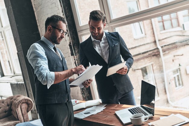 Team di innovatori. Due giovani uomini moderni in abiti da cerimonia che lavorano insieme stando in piedi in casa