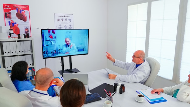 Team di personale medico che discute durante la videoconferenza con il medico nell'ufficio dell'ospedale. Medici che tengono riunioni online nella sala riunioni con uno specialista, infermiere che prendono appunti negli appunti.