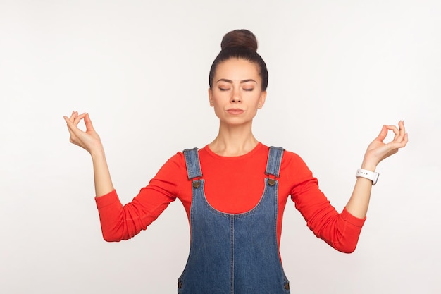 Tecnica del respiro dell'equilibrio mentale Ritratto di calma ragazza carina ed elegante con crocchia per capelli in tuta di jeans che si tiene per mano nel gesto di mudra e meditando praticando yoga indoor girato in studio su sfondo bianco