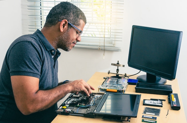 Tecnico informatico con gli occhiali, fissando il computer portatile sulla scrivania, con gli strumenti intorno. Uomo nero. Attrezzi da lavoro.