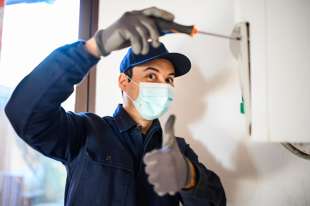 Tecnico sorridente che ripara uno scaldabagno indossando una maschera, concetto di coronavirus