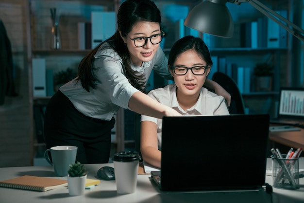 tecnologia di scadenza e concetto di persone. team aziendale di avvio creativo con computer portatile che lavora insieme in ufficio a tarda notte. due giovani colleghe asiatiche che parlano punto sullo schermo sorridendo.