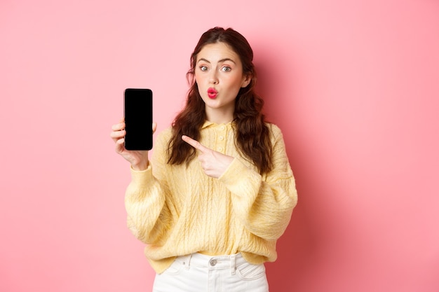 Tecnologia e acquisti online. La ragazza eccitata sembra curiosa, punta il dito sullo schermo del cellulare vuoto, mostra l'app sullo smartphone, in piedi contro il muro rosa.