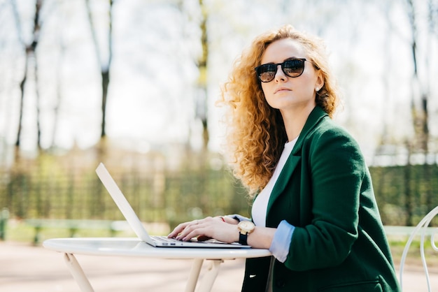 Tecnologia e comunicazione per il tempo libero delle persone Bella donna d'affari che indossa occhiali da sole e giacca elegante utilizzando il computer portatile per conversare con i suoi partner seduti lateralmente alla scrivania bianca