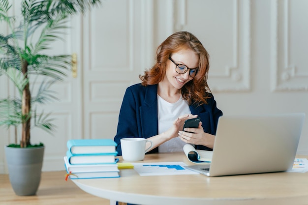 Tecnologia e concetto di comunicazione La giovane femmina dai capelli rossi controlla la casella di posta elettronica sul telefono cellulare moderno ha un sorriso piacevole studia i documenti possiede una grande azienda utilizza il computer portatile per lavoro