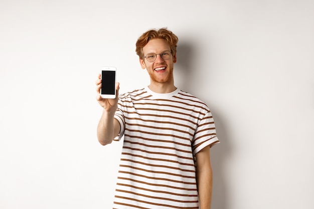 Tecnologia e concetto di e-commerce. Felice giovane uomo dai capelli rossi con gli occhiali che mostra lo schermo vuoto dello smartphone, sorridendo soddisfatto, in piedi su sfondo bianco.