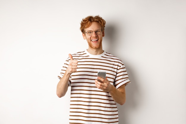 Tecnologia e concetto di e-commerce. Modello maschio soddisfatto con capelli rossi, mostrando il pollice in su e tenendo lo smartphone, sfondo bianco.