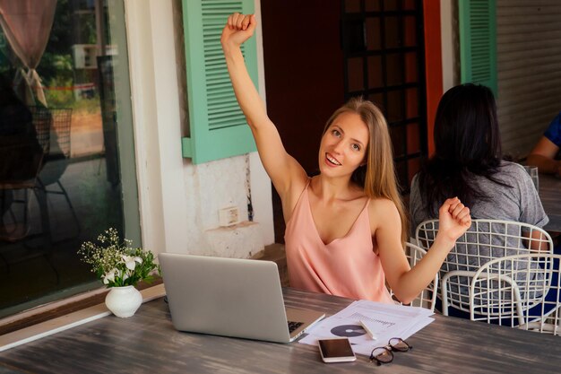 Tecnologia e viaggi. Lavorare all'aperto. Concetto di libero professionista. Piuttosto giovane donna che utilizza computer portatile nella caffetteria sulla spiaggia tropicale. Evviva il successo della vittoria e la promozione del concetto di affare di successo al lavoro