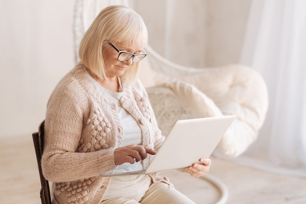 Tecnologia moderna. Piacevole bella donna invecchiata che tiene un laptop e guarda il suo schermo mentre era seduto nella sua stanza