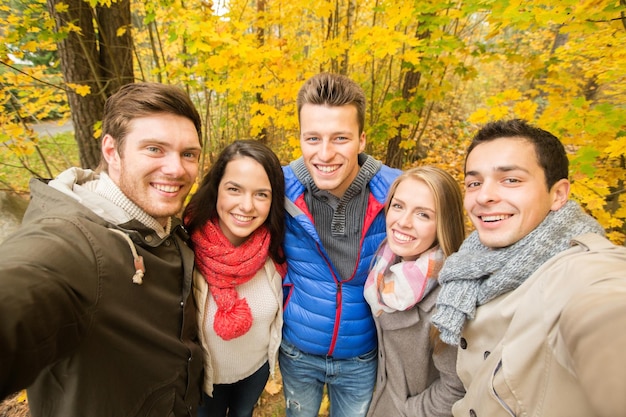 tecnologia, stagione, amicizia e concetto di persone - gruppo di uomini e donne sorridenti che prendono selfie con smartphone o fotocamera nel parco autunnale