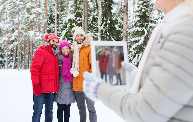 tecnologia, stagione, amicizia e concetto di persone - gruppo di uomini e donne sorridenti che scattano foto con computer tablet pc nella foresta invernale