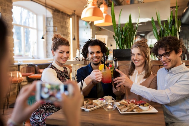 tecnologia, stile di vita, vacanze e concetto di persone: amici felici che tintinnano bevande e fotografano tramite smartphone al bar o al caffè