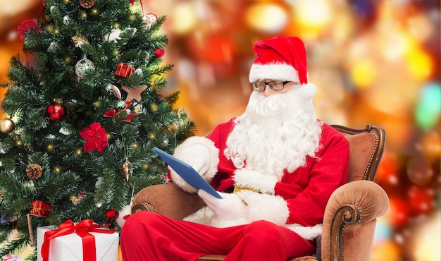 tecnologia, vacanze e concetto di persone - uomo in costume di babbo natale con computer tablet pc, regali e albero di natale seduto in poltrona su sfondo a luci rosse