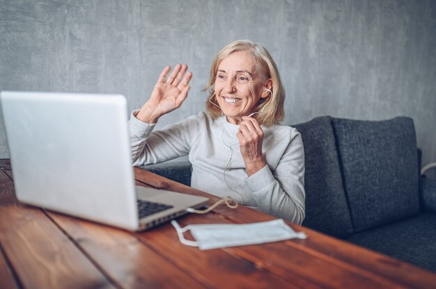 Tecnologia, vecchiaia e concetto della gente - donna senior più anziana felice con la maschera medica del fronte che lavora e che fa una video chiamata con il computer portatile a casa durante la pandemia del coronavirus COVID19. Stare a casa