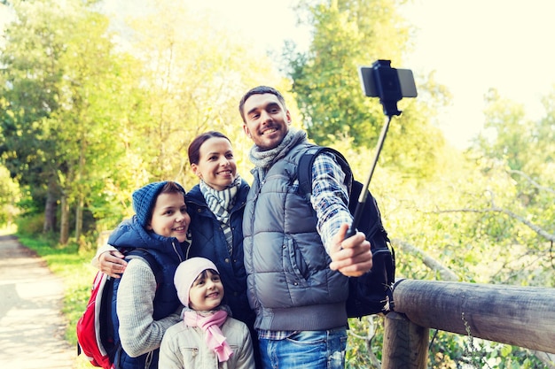 tecnologia, viaggi, turismo, escursione e concetto di persone - famiglia felice con zaini che scattano foto con lo smartphone su selfie stick ed escursioni