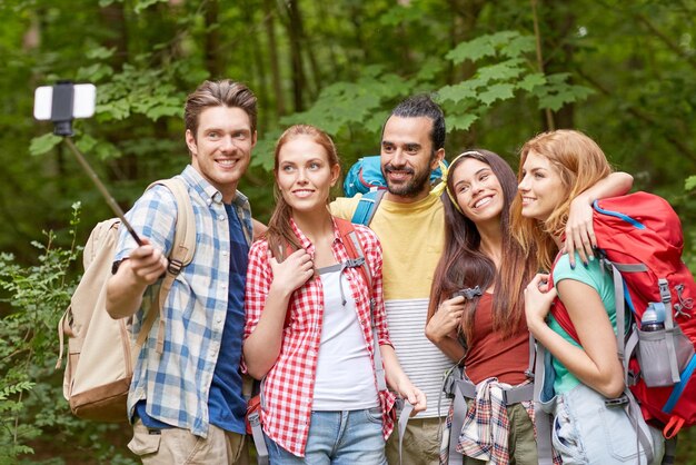 tecnologia, viaggi, turismo, escursione e concetto di persone - gruppo di amici sorridenti che camminano con gli zaini per scattare foto con lo smartphone sul selfie stick nei boschi