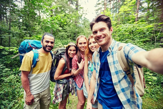 tecnologia, viaggi, turismo, escursione e concetto di persone - gruppo di amici sorridenti che camminano con gli zaini prendendo selfie con smartphone o fotocamera nei boschi