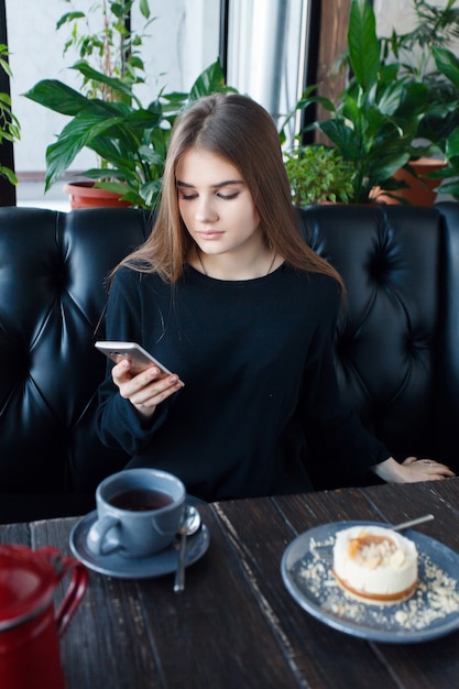 Tecnologie, emozioni, stile di vita, persone, concetto di adolescenti - Giovane donna felice che legge sul suo telefono cellulare mentre è seduta in un moderno interno della caffetteria, splendida ragazza hipster con un bel sorriso