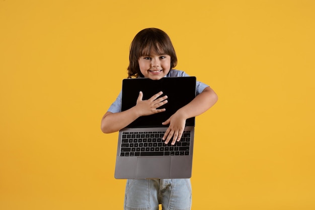 Tecnologie moderne e bambini Simpatico ragazzino che porta un laptop aperto e sorride allo sfondo giallo dello studio della fotocamera