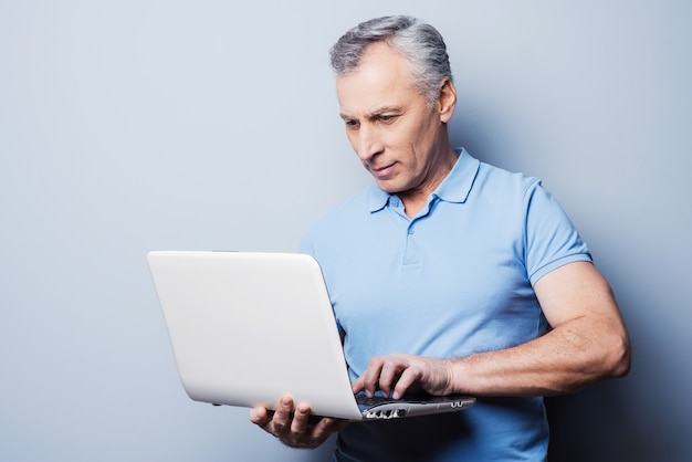 Tecnologie moderne sempre a portata di mano. Uomo anziano allegro in computer portatile della tenuta casuale mentre stando contro il fondo grigio