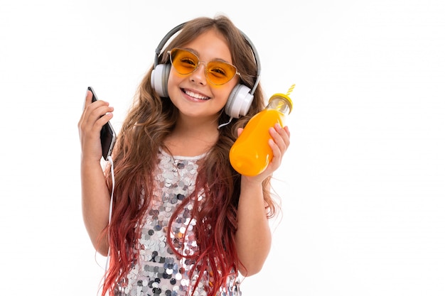 Teen donna con lunghi capelli biondi tinti di punte rosa, in abito chiaro lucido, scarpe da ginnastica bianche e nere, in piedi con le cuffie, con telefono e succo in bottiglia di vetro con tubo a strisce in mano