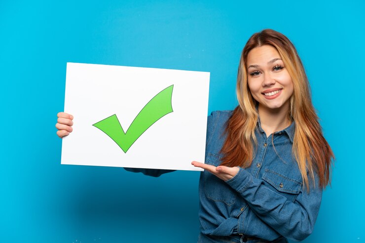 Photo teenager girl over isolated blue background holding a placard with text green check mark icon and  pointing it