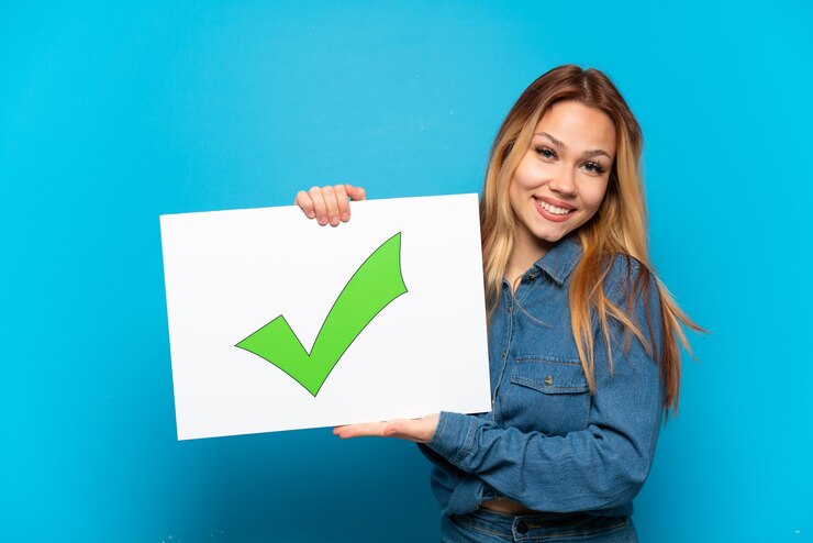 Photo teenager girl over isolated blue background holding a placard with text green check mark icon with happy expression