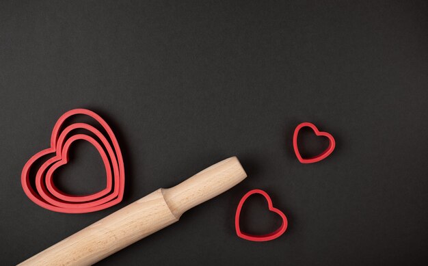 Teglie a forma di cuore rosse per biscotti e mattarello di legno su sfondo scuro (nero). Concetto di San Valentino.