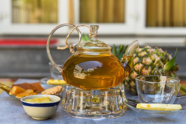 Teiera di vetro con tè all'ananas e bicchieri di marmellata fatti in casa e frutta cristallizzata su sfondo scuro