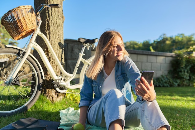 Telecomunicazioni videochiamata videoconferenza comunicazione con amici e familiari a distanza Adolescente studentessa che guarda lo schermo dello smartphone ridendo parlando seduto sull'erba nel parco