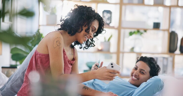 Telefonare a una coppia di lesbiche e ridere di meme sui social media o video divertenti nel salotto della loro casa ragazze LGBT felici e legami con scherzi sullo smartphone mentre si rilassano sul divano nel salotto per la felicità