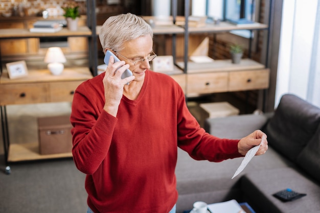 Telefonata. Uomo anziano intelligente serio che tiene il suo telefono cellulare e che effettua una chiamata mentre guarda le note