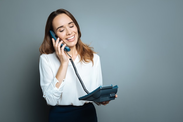 Telefoni con filo. Piacevole donna graziosa deliziata che tiene un telefono con filo e si mette il ricevitore all'orecchio mentre effettua una chiamata