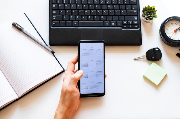 Telefono cellulare con calendario aperto per l'anno 2022 in mano maschile su scrivania, laptop, penna, orologio, chiavi