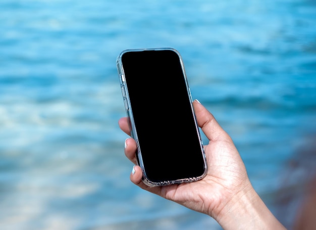 Telefono cellulare da spiaggia con schermo vuoto Immagine mockup dello schermo del desktop vuoto nero in mano che tiene in mano uno smartphone mobile sullo sfondo blu del mare in estate Prenotazione di viaggi alla ricerca di marketing aziendale