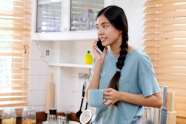 Telefono di conversazione della giovane donna asiatica adolescente in cucina, tazza di caffè asiatica della tenuta della ragazza mentre Smart Phone di conversazione con stile di vita di felicità di mattina, della gente e di tecnologia