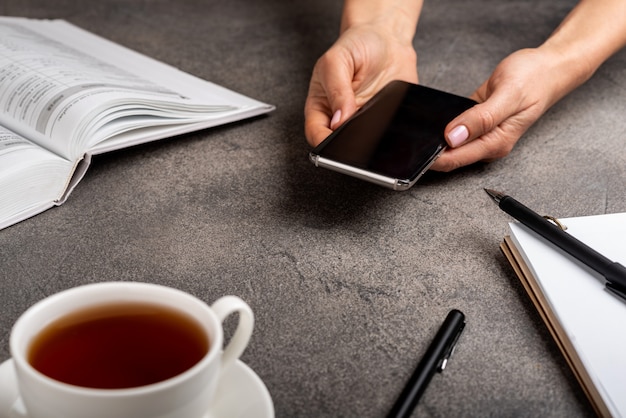 Telefono nelle mani della donna con libro, taccuino, penne e tazza di tè