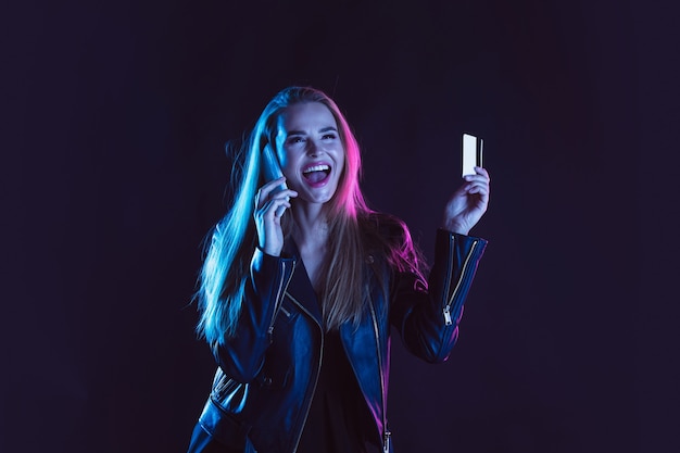 Telefono parlante, felice. Ritratto di giovane bella donna alla luce al neon su sfondo scuro studio. Le emozioni umane, venerdì nero, lunedì cibernetico, acquisti, vendite, concetto di finanza. Copyspace.