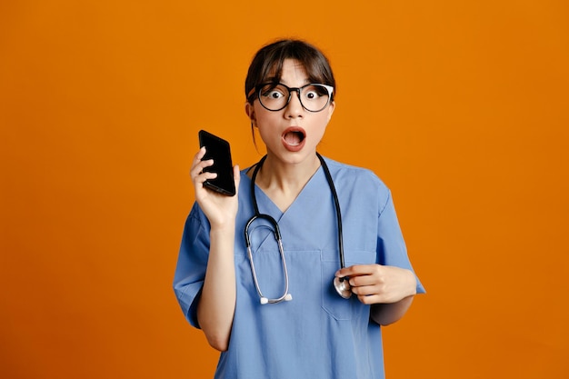 telefono spaventato giovane dottoressa che indossa uno stetoscopio fitth uniforme isolato su sfondo arancione