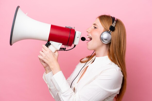 Telemarketer donna rossa che lavora con un auricolare isolato su sfondo rosa che grida attraverso un megafono