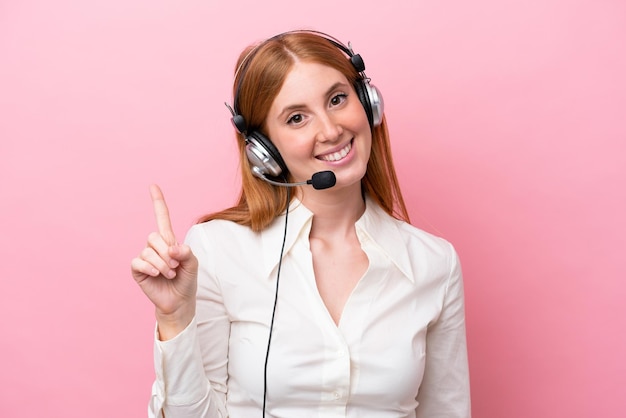 Telemarketer donna rossa che lavora con un auricolare isolato su sfondo rosa che mostra e alza un dito in segno di meglio