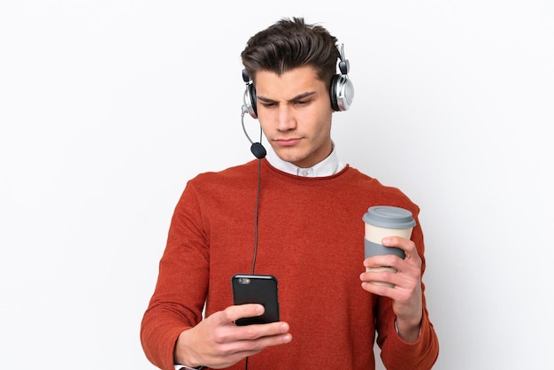 Telemarketer uomo caucasico che lavora con un auricolare isolato su sfondo bianco tenendo il caffè da portare via e un cellulare