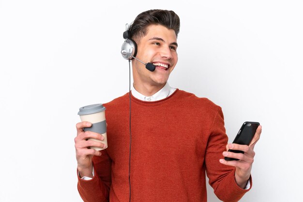 Telemarketer uomo caucasico che lavora con un auricolare isolato su sfondo bianco tenendo il caffè da portare via e un cellulare