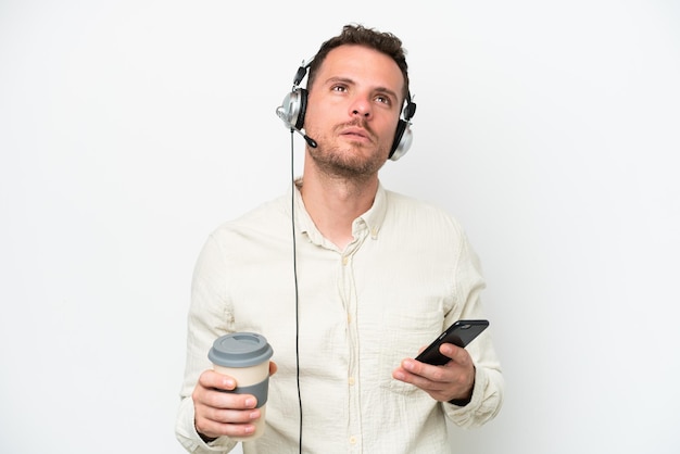 Telemarketer uomo caucasico che lavora con un auricolare isolato su sfondo bianco tenendo il caffè da portare via e un cellulare