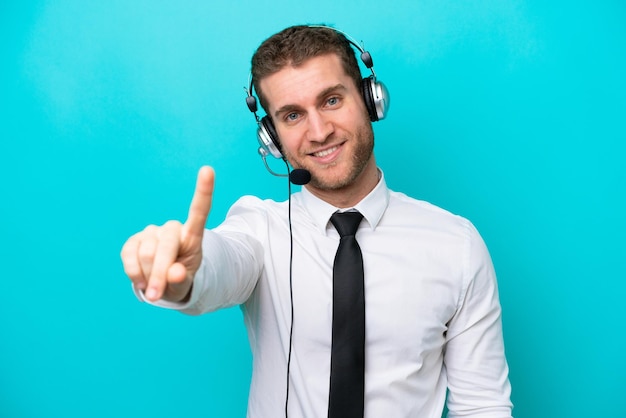 Telemarketer uomo caucasico che lavora con un auricolare isolato su sfondo blu che mostra e solleva un dito