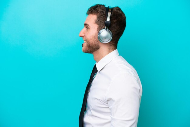 Telemarketer uomo caucasico che lavora con un auricolare isolato su sfondo blu ridendo in posizione laterale